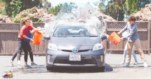A group of people working together, playfully splashing water on a car with buckets during a sunny outdoor car wash.