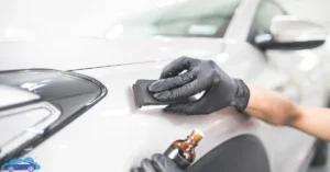 A professional applying ceramic coating to a white car's surface using a sponge applicator while wearing black gloves.